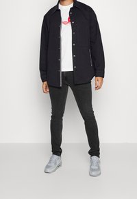 Chaqueta negra con cierres de botón sobre una camiseta blanca con un gráfico rojo. Jeans grises y zapatillas gris claro completan el atuendo.