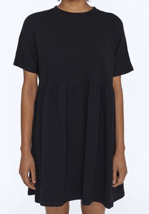 Kobieta ubrana w luźną, czarną sukienkę z krótkim rękawem, ze ściąganym w talii i okrągłym dekoltem, stojąca na jednolitym tle.