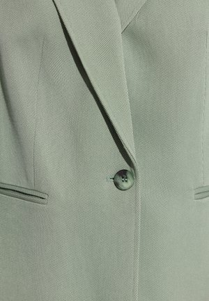 Zbliżenie jasnobłękitnej marynarki z wyciętym klapem, jednym ciemnym guzikiem i dwoma poziomymi kieszeniami z rozcięciem.