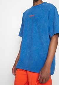 Blå oversized t-shirt med en urtvättad struktur och rund halsringning, med röd text "LES ENFANTS" på bröstet. Matchas med oranga byxor.