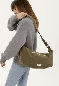 Sac à main vert olive en toile avec une forme incurvée, doté d'une poche zippée et d'une sangle réglable.