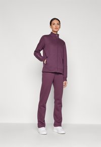 Fioletowy zestaw bluzy i spodni, wykonany z miękkiego materiału. Kurtka ma wysoki kołnierz, kieszenie na zamek i boczne pasy. Ubranie ma luźny krój.
