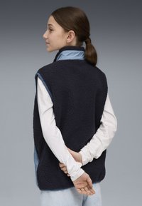 Navy Fleece-Weste mit hohem Kragen, strukturiertem Äußeren und blauer Innenausstattung. Getragen über einem weißen Langarmshirt, die Hände hinter dem Rücken verschränkt.