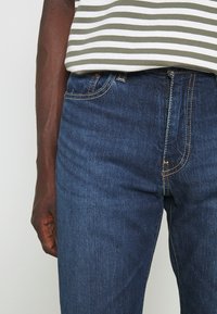 Dark blue denim jeans with yellow stitching, featuring a fitted design and a front pocket. Worn with a striped olive green top.