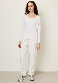 Noor naine lokkis juustega, seljas lahtine valge pikkade varrukatega särk, valged püksid ning hallid ja rohelised spordijalatsid, seisab valge seina ees.