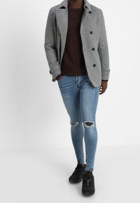 Grijze wollen dubbele jacks, bordeauxrode gebreide trui, versleten lichtblauwe skinny jeans en zwarte sneakers.