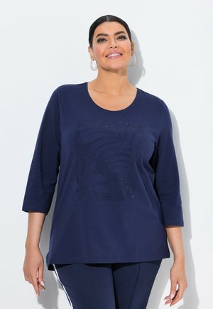 Lächelnde Frau mit dunklen Haaren trägt ein marineblaues Oberteil mit 3/4-Ärmeln und einem dezenten Blattmuster, kombiniert mit marineblauen Hosen, die weiße Seitenstreifen aufweisen.