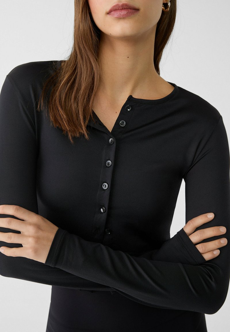 Haut noir à manches longues en tissu lisse, avec un col henley à boutons et des poignets ajustés avec des trous pour les pouces. Design simple et épuré.
