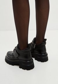Bottes chelsea en cuir noir avec surface texturée, semelle épaisse et fermeture éclair sur le côté. Possède une sangle Velcro à l'avant.
