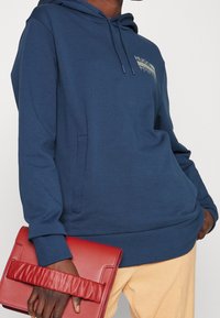 Felpa blu navy con tasca a marsupio e cappuccio con cordino, abbinata a una pochette in pelle rossa con manico arricciato.