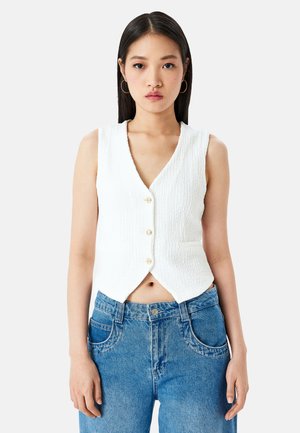 Canottiera bianca senza maniche, corta, realizzata in tessuto strutturato, con scollatura a V e cinque bottoni sul davanti. Indossata con jeans in denim blu.
