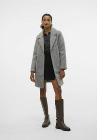 Manteau gris texturé avec revers à cran, associé à une jupe noire et des bottes en cuir marron jusqu'au genou. Ongles peints en rouge visibles.