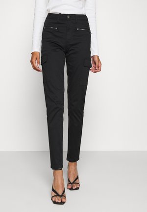 Schwarz, schlank geschnittene Cargo-Hose mit Reißverschlusstaschen vorne, kombiniert mit einem weißen Langarmshirt und schwarzen, geschlossenen Sandaletten mit Absatz.