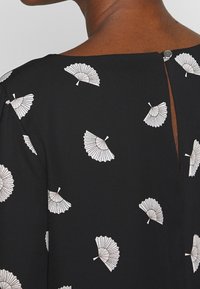 Svart klänning med vita fläktmönster, en nyckelhålsdetalj i ryggen och en knappstängning vid halsen. Slät tyg med en svag lyster.