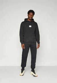 Felpa nera con cappuccio, dotata di tasca frontale e patch con logo, abbinata a pantaloni della tuta neri. Il modello indossa sneaker bianche e nere.