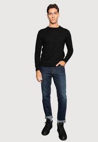 Hombre vestido con un suéter negro de manga larga, jeans oscuros arremangados y zapatillas negras, de pie con una mano en el bolsillo.