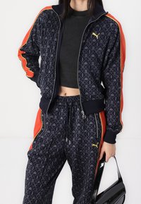 Navy tracksuit with geometric pattern, orange and yellow accents. Features zip front jacket and drawstring pants, paired with a black bag.