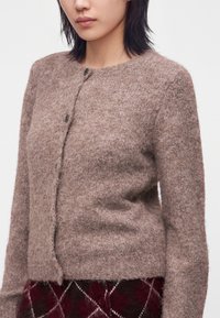 Young woman wearing a soft, fuzzy light brown buttoned cardigan and red and black plaid skirt against a plain white background.