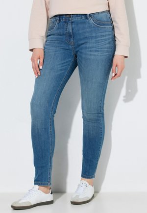 Person iført lyseblå skinny jeans, beige langærmet top og hvide sneakers, der står mod en hvid baggrund.