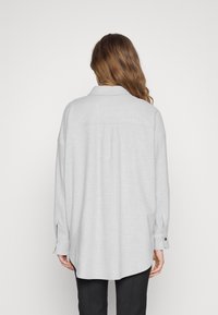 Camisa de manga larga gris claro, con cuello, puños abotonados y corte relajado. Tela texturizada y detalle de pliegue en la parte posterior.