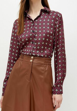Woman wearing a long-sleeve patterned blouse with pink, white, and brown geometric shapes tucked into a high-waisted brown leather skirt.