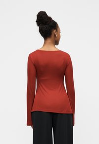 Μακρυμάνικο, εφαρμοστό τοπ σε σκούρο κόκκινο, κατασκευασμένο από απαλό, ελαστικό ύφασμα. Διαθέτει φαρδιά λαιμόκοψη και στρογγυλεμένο τελείωμα. Φοριέται με μαύρο, άνετο παντελόνι.