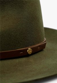 Sombrero de fieltro verde oliva con ala ancha, que presenta una banda de cuero marrón adornada con un emblema de calavera en tono dorado y costuras decorativas.