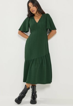 Femme portant une robe midi vert foncé avec des manches courtes volantées et des bottes noires à boucles mi-mollet, debout sur un fond blanc uni.