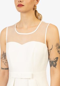 Abito bianco con una scollatura trasparente, scollo rotondo e corsetto aderente. Presenta una cintura a fiocco e una gonna plissettata. Orecchini a cerchio dorati completano il look.