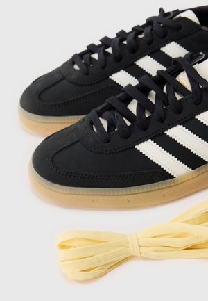 Baskets noires avec des bandes blanches Adidas et des semelles en gomme, à côté d'un rouleau de lacets jaune pâle sur une surface blanche.