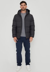 Sort polstret jakke med hætte, udstyret med lynlåslukning, båret over en grå skjorte og parret med navy blå bukser og hvide sneakers.