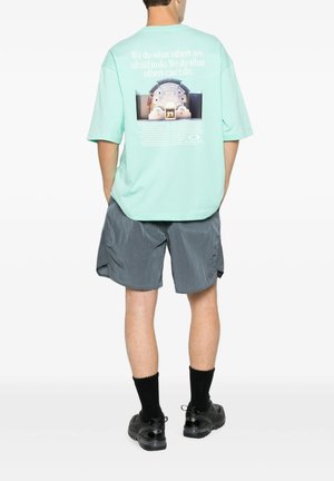 Uomo che indossa una maglietta oversize verde menta con testo e immagine stampati sulla schiena, pantaloncini grigi, calzini neri e scarpe da ginnastica nere, in piedi di spalle.