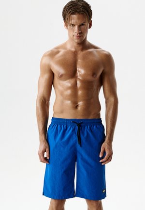 Muskularny mężczyzna bez koszulki w niebieskich kąpielówkach z czarnym sznurkiem, stojący na jednolitym białym tle.