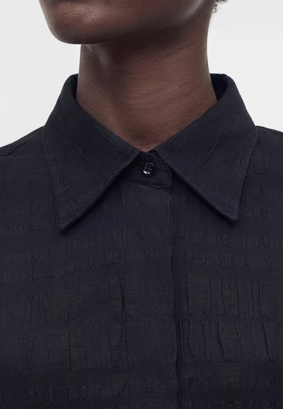 Κοντινό πλάνο σκοτεινόχρωμου ατόμου που φοράει μαύρο υφασμάτινο πουκάμισο με μυτερό γιακά, που κλείνει με ένα κουμπί.