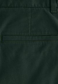 Dunkelgrüner Stoff von Hosen mit glatter Textur. Verfügt über eine Gesäßtasche mit einer horizontalen Naht und kontrastierenden Nähdetails.