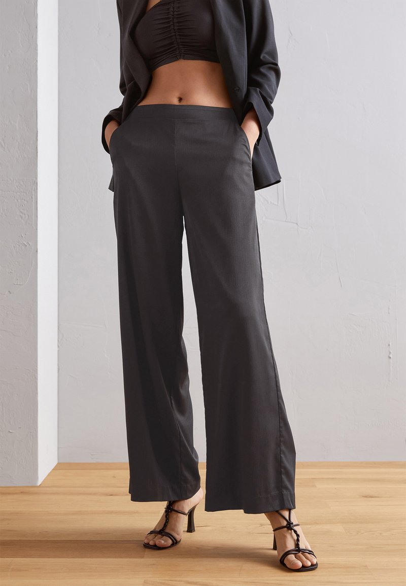 Pantalons gris à jambes larges avec une finition en tissu lisse, dotés de poches sur les côtés et d'une coupe ajustée. Associés à des sandales à talons hauts noires à lanières.