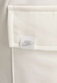 Bílá atletická tkanina sešitá s kapsičkou a malou šedou visačkou Nike. Textura vypadá hladce a jemně.