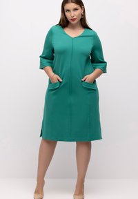 Vestido turquoise hasta la rodilla, hecho de tela suave, con escote en V, mangas de tres cuartos y bolsillos laterales. Textura lisa sin patrones.