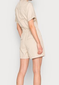 Beige krátký overal vyrobený z bavlny, s límečkem, krátkými rukávy, elastickým pasem a dvěma zadními kapsami.