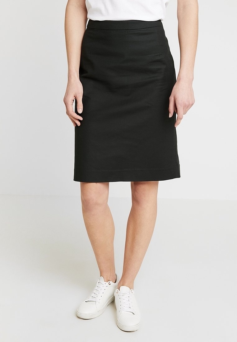 Femme portant une jupe noire longueur genou, un t-shirt blanc et des baskets blanches, debout devant un fond clair uni.