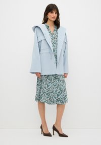 Abrigo azul claro de gran tamaño con capucha, grandes bolsillos frontales, sobre un vestido verde floral de manga corta y dobladillo con volantes, combinado con tacones marrones.