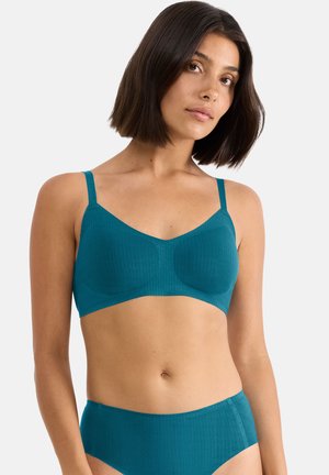 Vrouw met donker schouderlang haar, gekleed in een teal ribgebreide beha en bijpassende hoog uitgesneden slips, tegen een effen achtergrond.