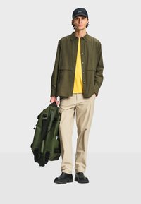 Homme portant une veste verte boutonnée, une chemise jaune, un pantalon beige, des chaussures noires et une casquette noire, tenant un grand sac de voyage vert olive.