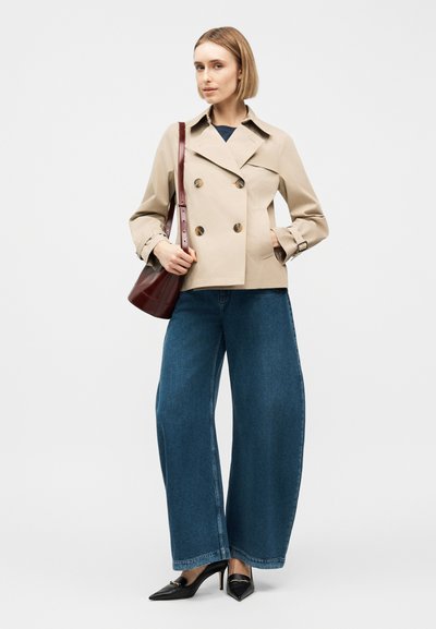 Femme portant une veste beige à double boutonnage, un jean bleu à jambes larges, des talons noirs, et tenant un sac à bandoulière marron foncé, debout devant un fond blanc.