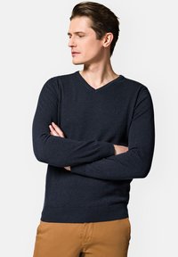 Ciemnoniebieski sweter w serek z dzianiny, z ribbowymi mankietami i dołem, z małym logo na klatce piersiowej. W zestawie z beżowymi spodniami.