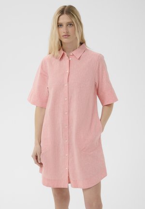 Femme debout portant une robe chemise rose clair à manches courtes, boutonnée, avec un col et deux poches avant.