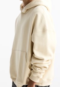 Béžová mikina s kapucí, přední kapsou, volným střihem a nastavitelnou šňůrkou na kapuci. Vybavena žebrovanými manžetami a jemným logem na rukávu.