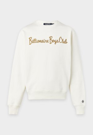 Giovane uomo che indossa una felpa color crema con la scritta "Billionaire Boys Club" e pantaloni sportivi verde scuro con sneakers bianche su uno sfondo semplice.