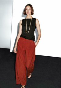Black sleeveless top, loose reddish-brown wide-leg pants, and a light bead necklace. Model stands on a dark floor against a white backdrop.