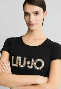 Juoda trumpų Rankovių T-shirt su "LIU:JO" logotipu gyvūnų raštu, papuoštas akmenukais. Minkšta medžiaga su apvaliu iškirpimu.